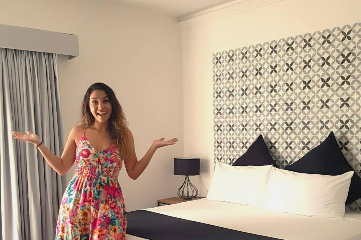 Une chambre d’hôtel avec un lit, une table de chevet, une lampe, une valise et une fenêtre avec des rideaux.