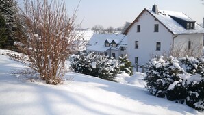 A snow-covered residential area with a white house featuring a steep roof and multiple windows.