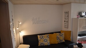 A modern living room with a black sofa, patterned cushions, a 'Welcome' sign, and a shelf with a printer.