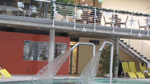 A swimming pool with two water jets, yellow lounge chairs, and a staircase leading to an upper deck with seating.