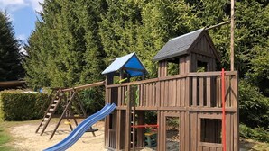 A wooden play structure with a slide and climbing elements, surrounded by sand and greenery.