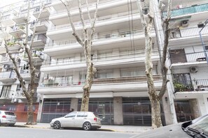 A multi-story residential building with balconies, a parked car, and trees in front.