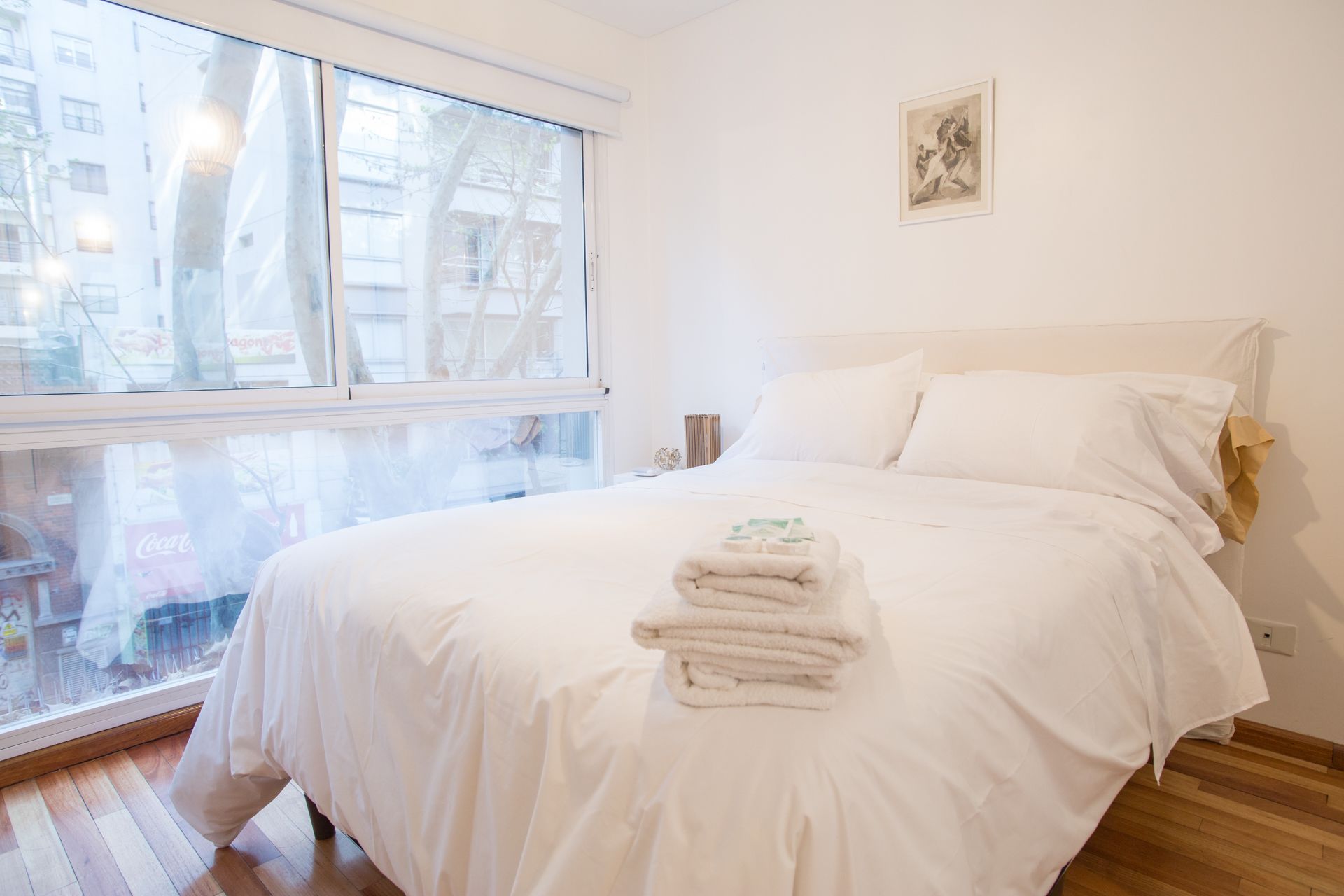 A hotel room with a large bed, a window with a view of buildings, and a framed picture on the wall.