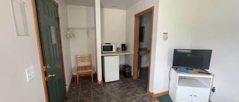 Una habitación pequeña con una puerta verde, un escritorio con un ordenador, un televisor sobre un mueble y una silla.