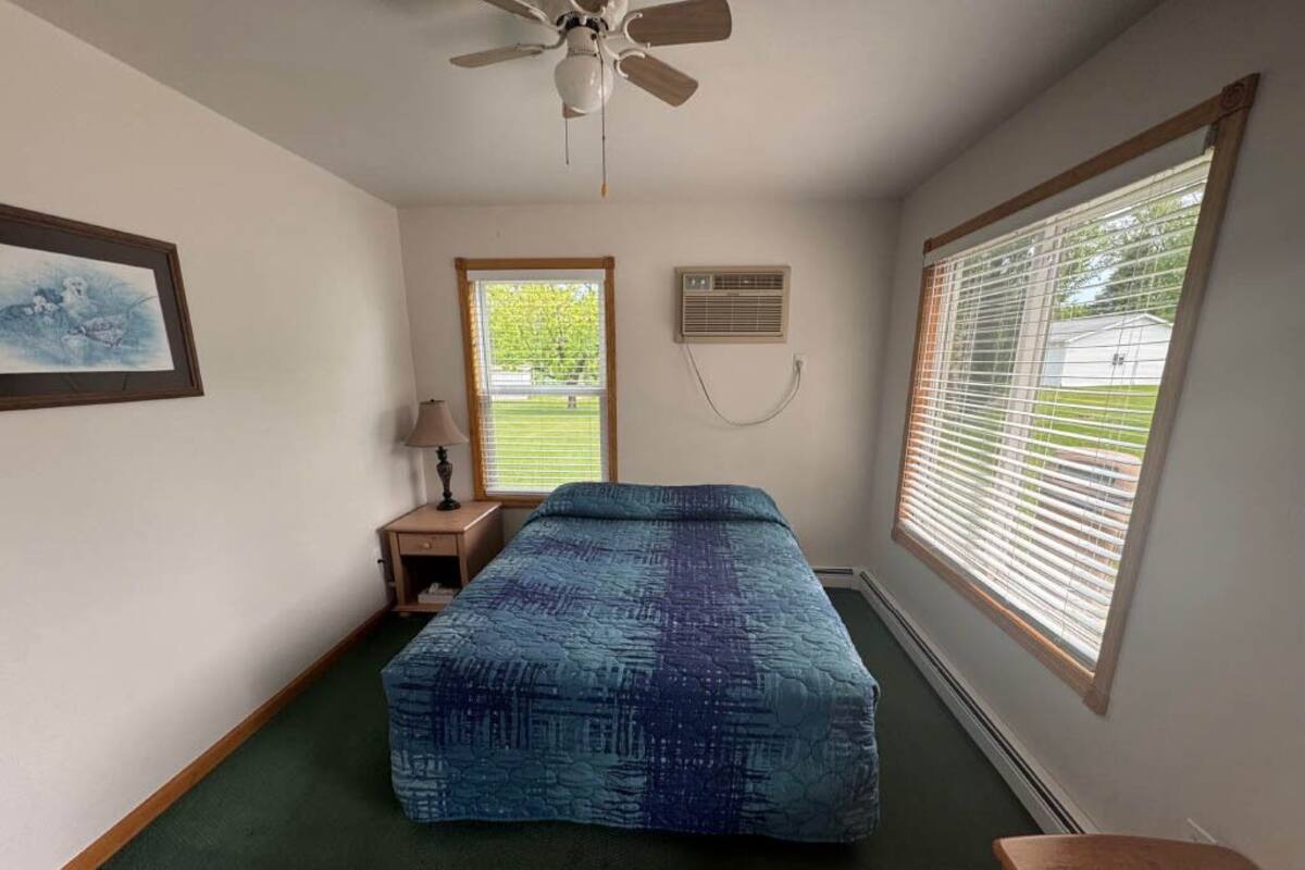 A bedroom with a bed, a nightstand, a lamp, a ceiling fan, and a window with blinds.