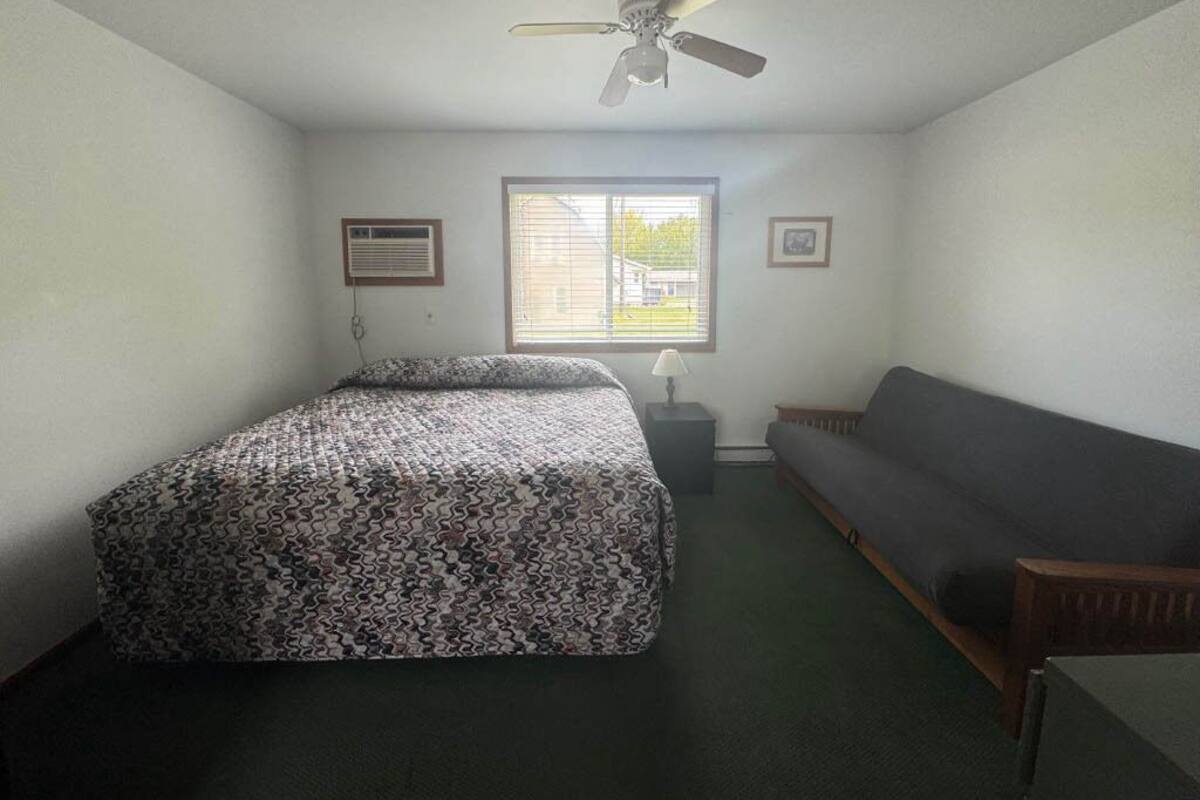 A small, sparsely furnished room with a bed, a sofa, a ceiling fan, and a window with blinds.