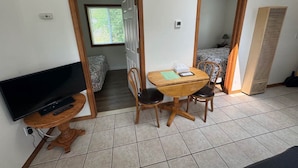 A compact room with a TV on a wooden stand, a small wooden table with two chairs, and a bed in the background.