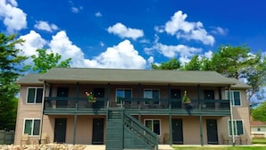 A two-story building with a balcony on the upper floor, a staircase leading to the entrance, and a clear blue sky with clouds.
