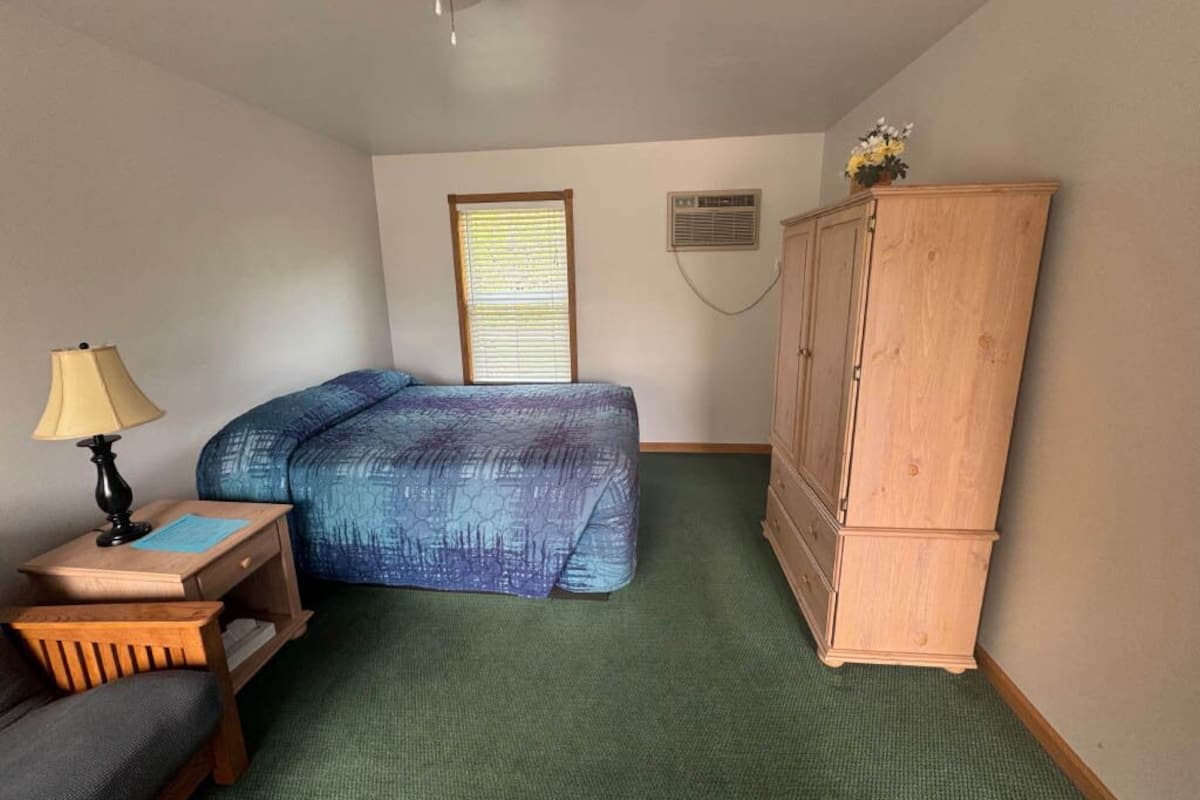 A small, neatly arranged room with a bed, a wooden cabinet, a desk with a lamp, and a window with blinds.