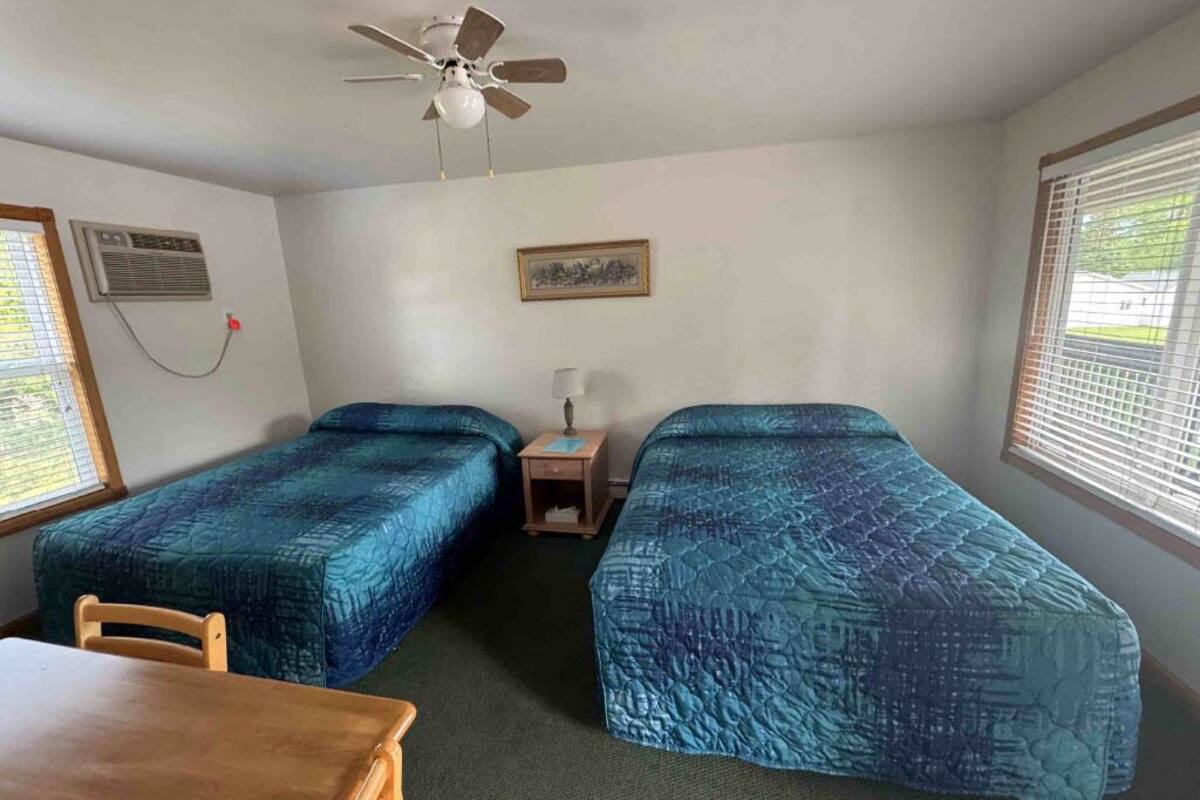 A room with two beds, a ceiling fan, a table, and a window with blinds.