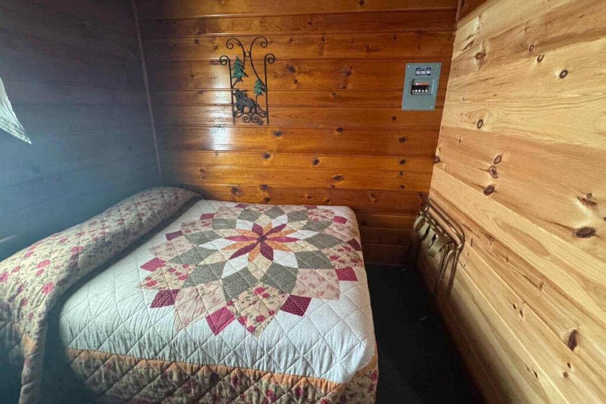 A small, cozy cabin room with a wooden interior, a bed with a quilt, and a wall-mounted thermostat.