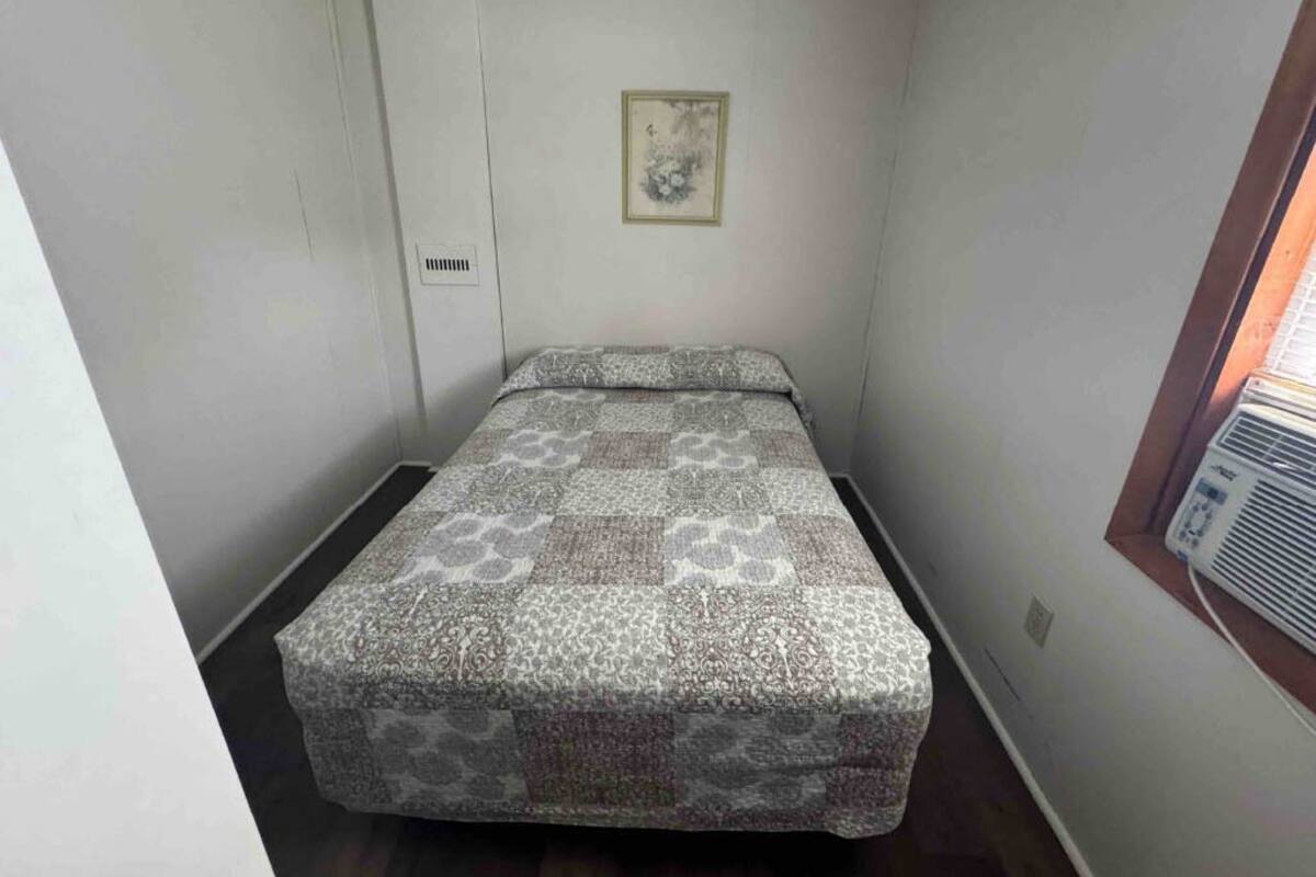A small, narrow room with a single bed, a window with an air conditioner, and a wall-mounted vent.
