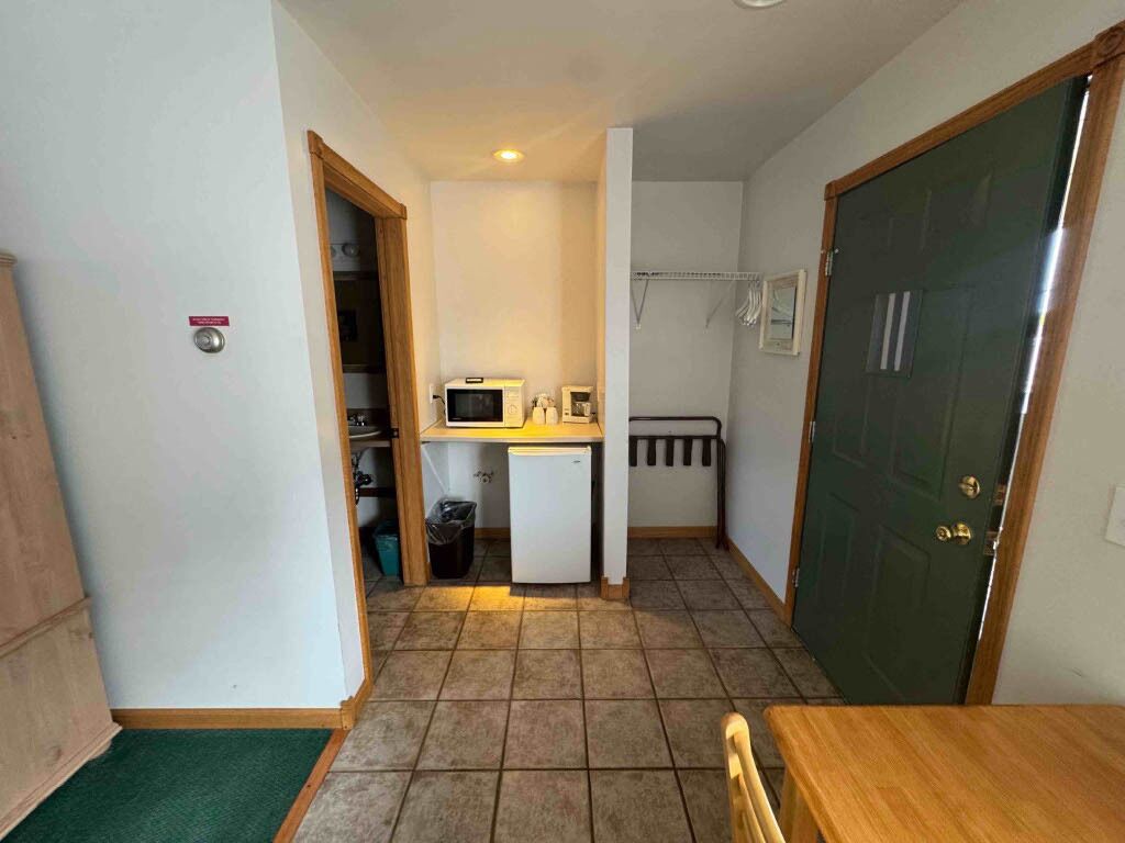 A compact kitchen with a microwave, small refrigerator, and a sink. There is a wooden table and chairs in the foreground.