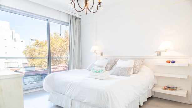 Ein modernes Schlafzimmer mit einem großen Bett, einer Kristallleuchter und Blick auf die Stadt durch große Fenster.