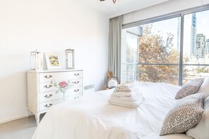 Een slaapkamer met een groot raam, een witte ladekast met bloemmotieven op de ladeknopjes, en een netjes opgemaakt bed met gevouwen handdoeken.