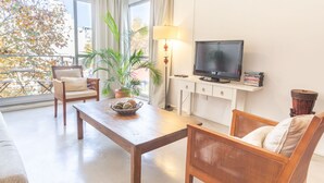 Ein Wohnzimmer mit Fernseher, einer Couch und Sesseln, einem Esstisch mit Obstschale und einer Topfpflanze.