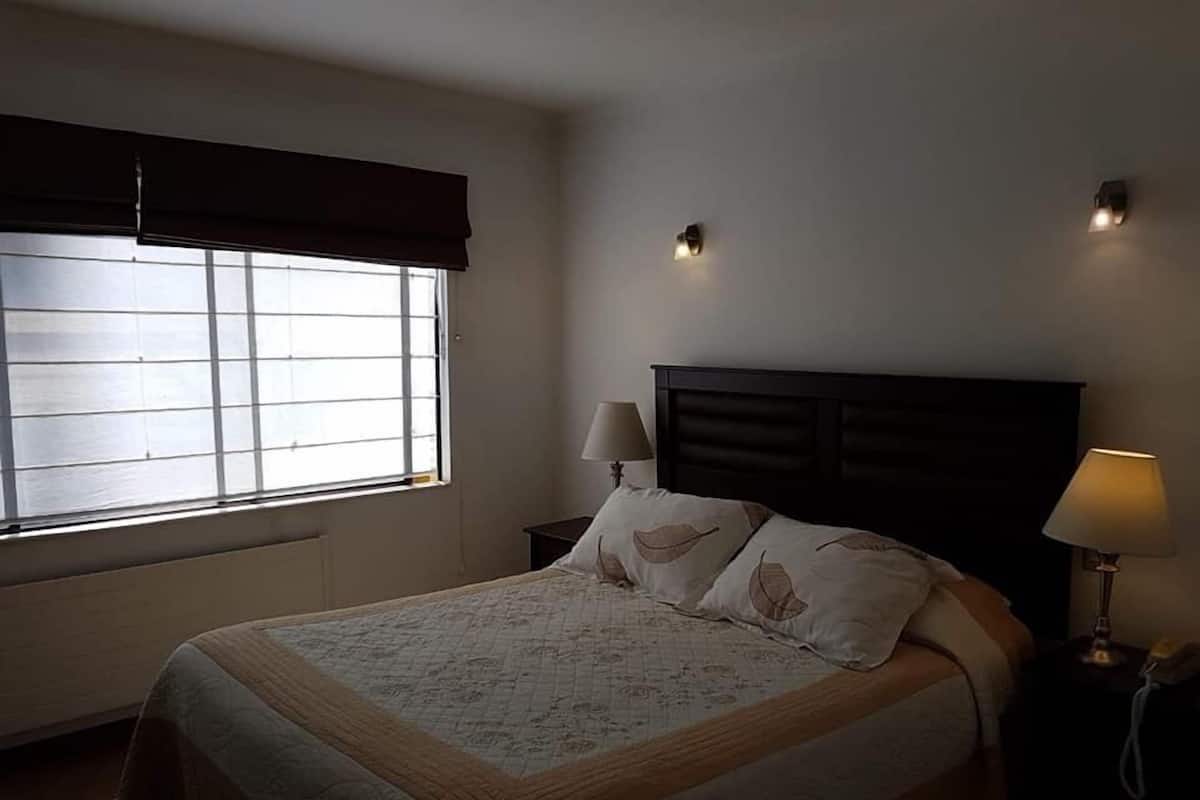 Un dormitorio con cama, dos lámparas de noche, una ventana con persianas y una mesita de noche con lámpara.