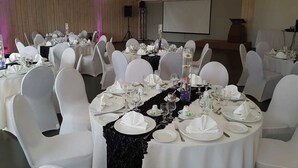 Un salón de banquetes con mesas redondas, sillas blancas y una pantalla para proyector en la pared.