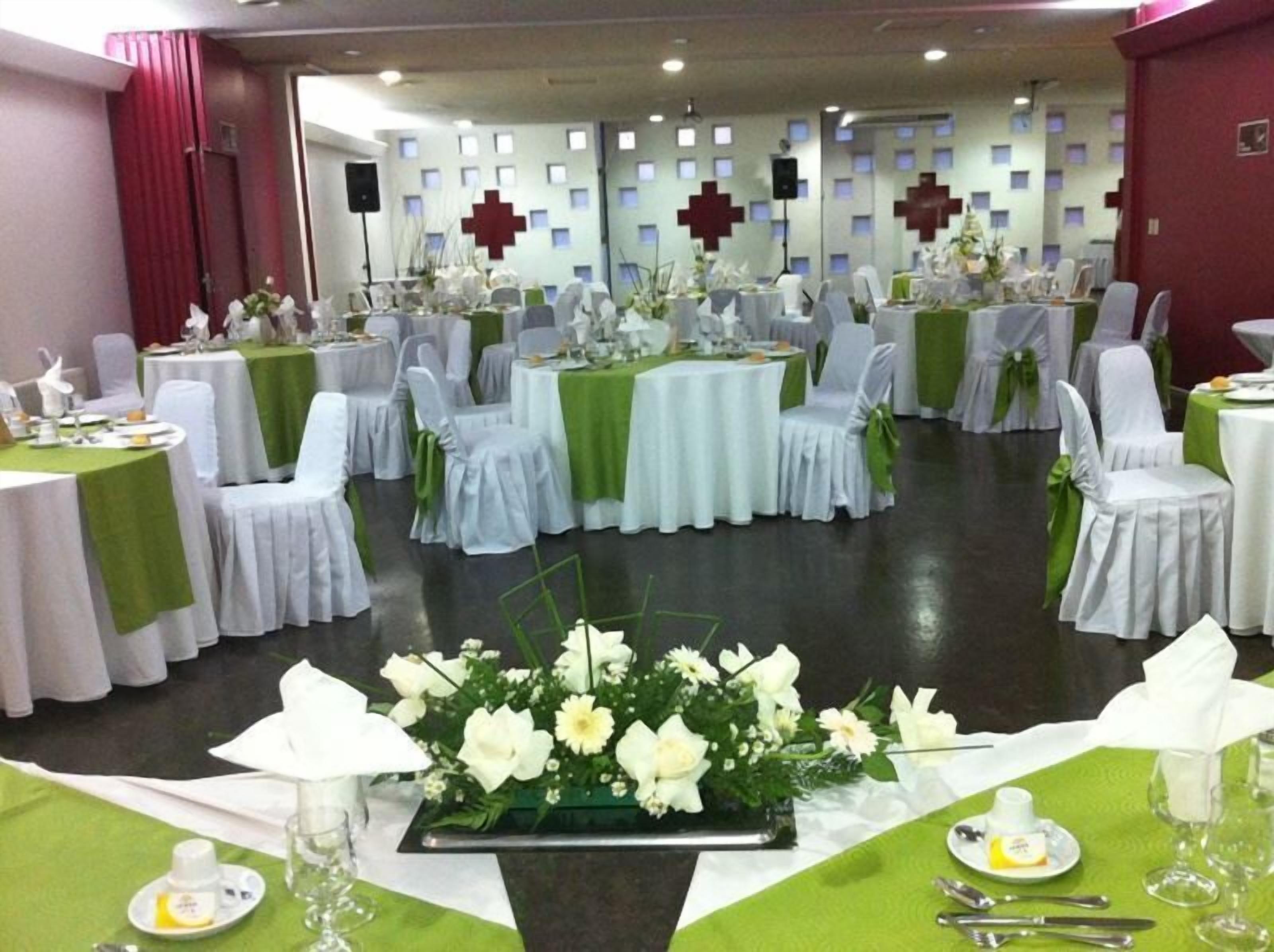 A banquet hall with round tables covered in white tablecloths and green sashes, chairs with white covers, and a floral centerpiece on each table.