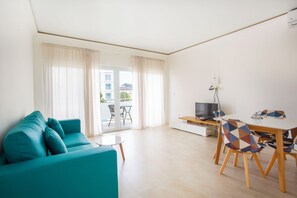 A modern living room with a teal sofa, a dining table with colorful chairs, a TV, and a sliding glass door leading to a balcony.