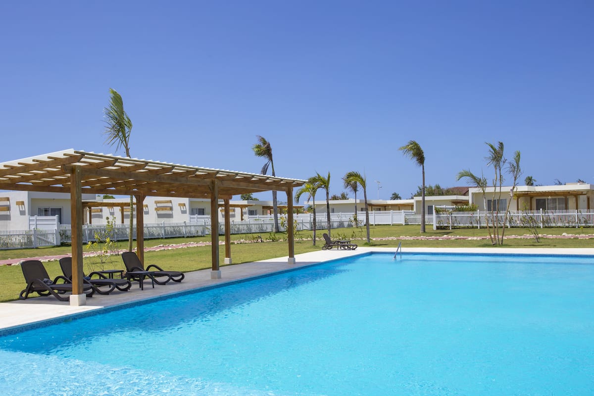 A swimming pool with lounge chairs and a wooden pergola, surrounded by palm trees and a white fence.