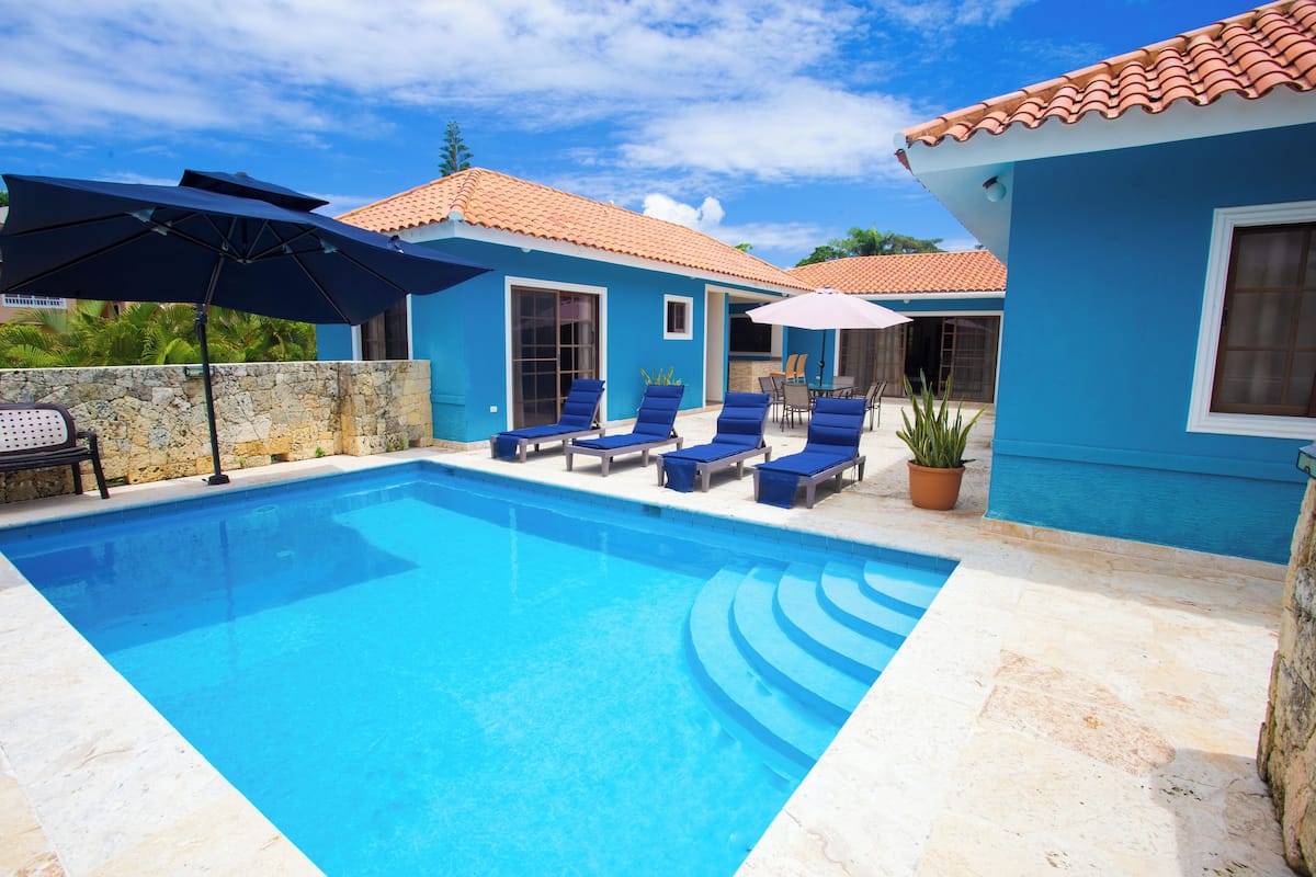 A swimming pool with blue lounge chairs and umbrellas, surrounded by a stone wall and a blue building with a tiled roof.