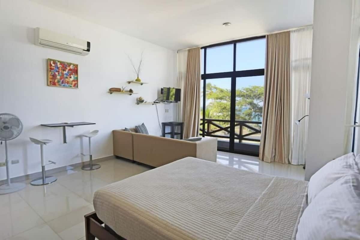 A hotel room with a bed, a sofa, a TV, a fan, and a balcony with a view.