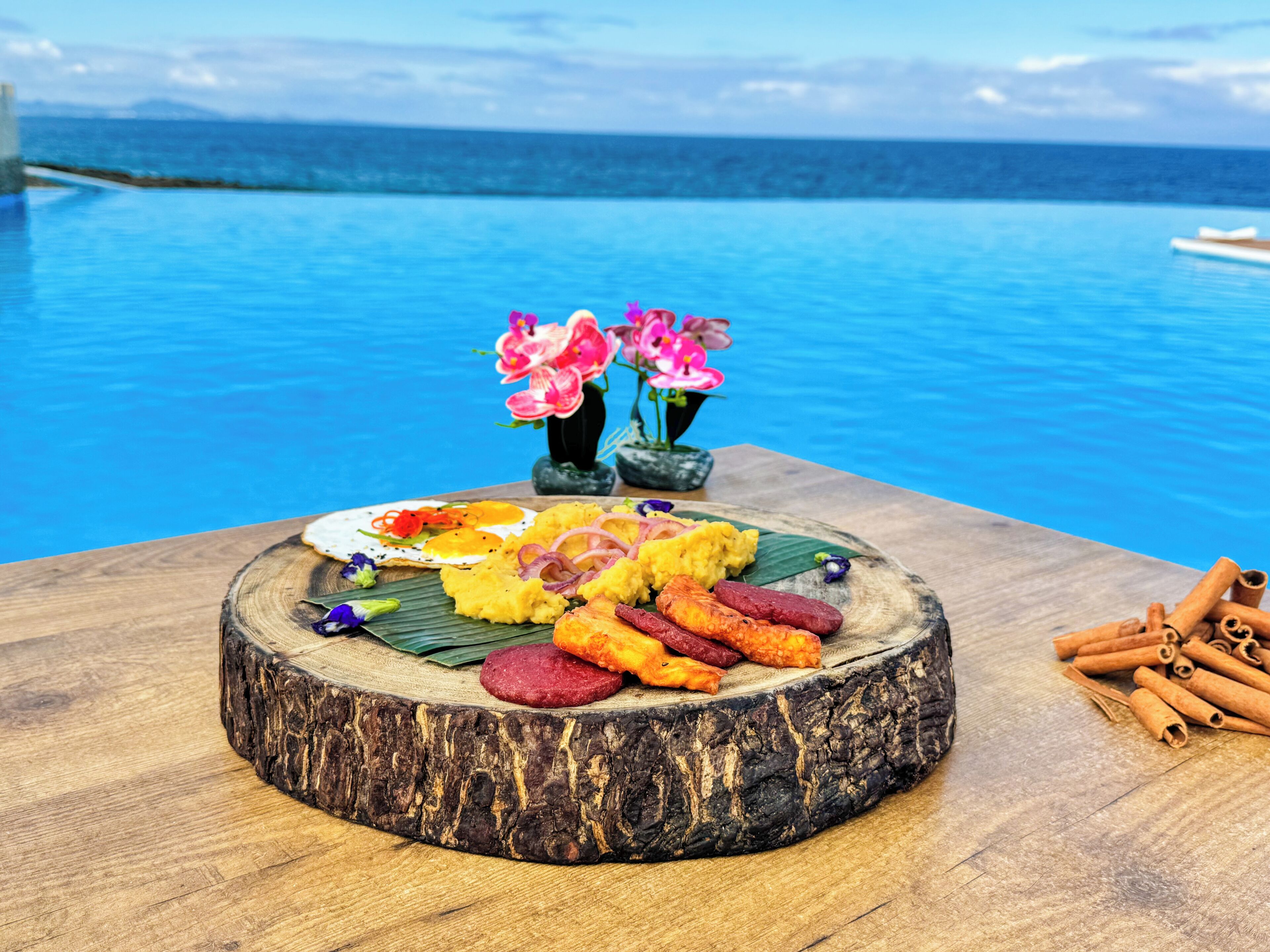 Een houten tafel met diverse soorten eten, waaronder eieren, worst en een banaan, met op de achtergrond een zwembad en uitzicht op zee.