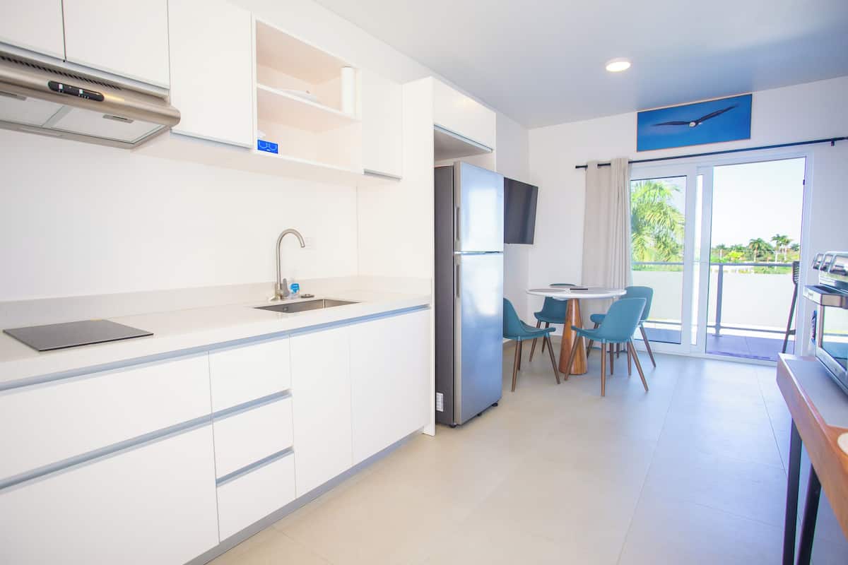 A modern kitchen with white cabinetry, a built-in stove, and a refrigerator. There is a dining area with a round table and blue chairs.