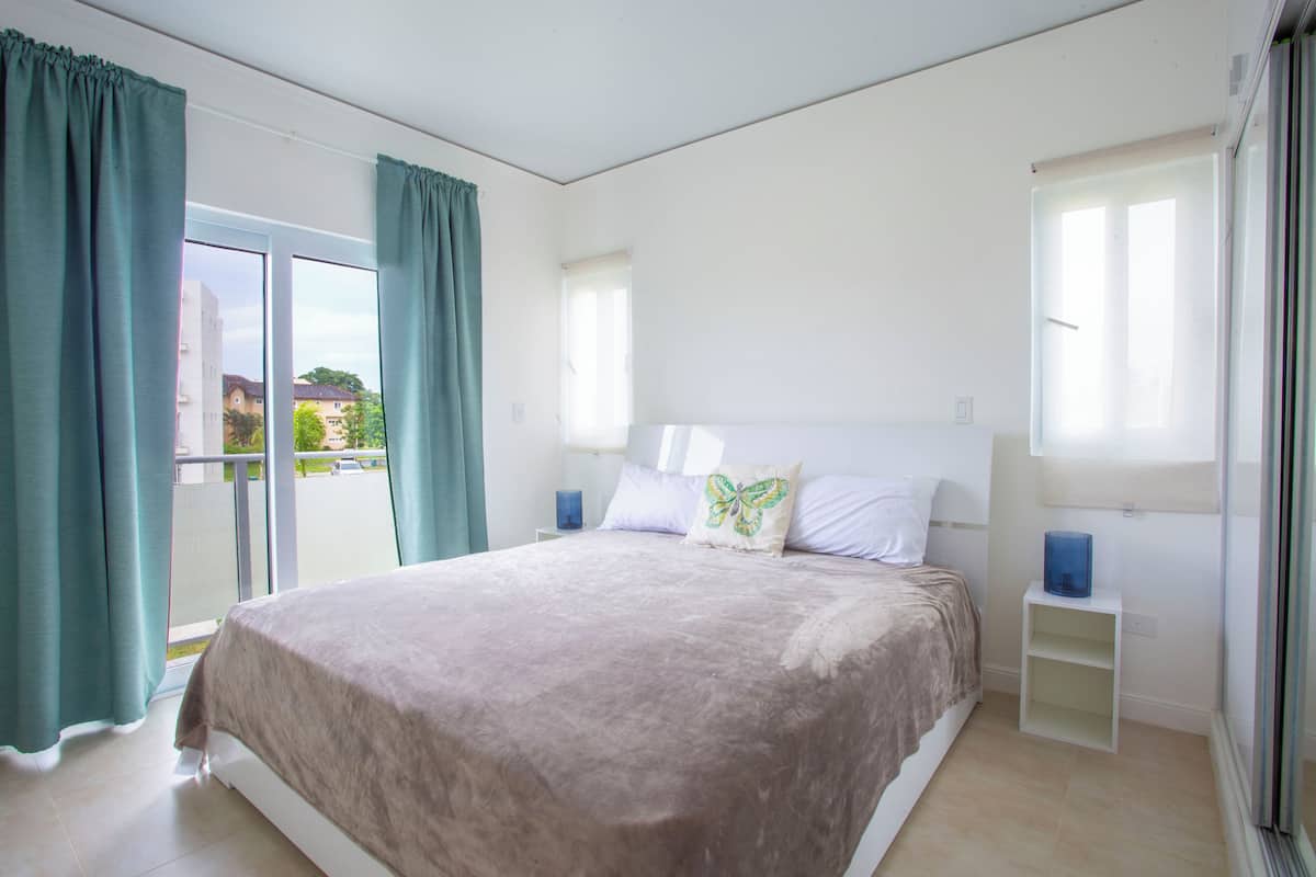 A bedroom with a large bed, a nightstand, a window with curtains, and a view of the outside.