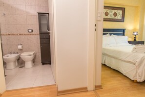 A hotel bathroom with a toilet, bidet, and cabinet, and a bedroom with a bed and nightstand.