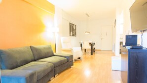 Een moderne woonkamer met een grijze bank, een eethoek met een tafel en stoelen, en een keuken met een computer en apparatuur.
