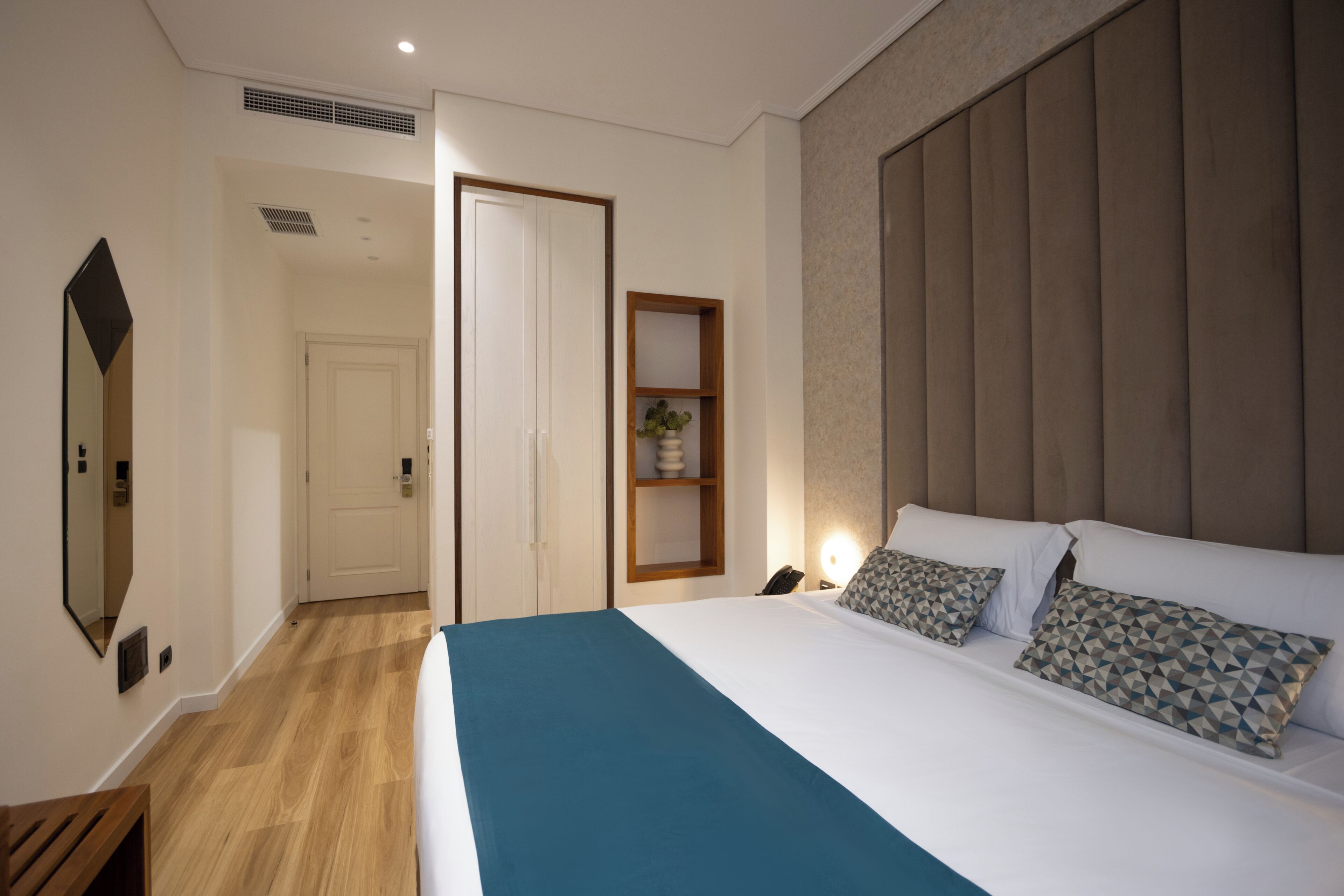 Une chambre d’hôtel comprenant un lit, une table de chevet, un miroir, une porte, une étagère avec une plante et un luminaire fixé au mur.