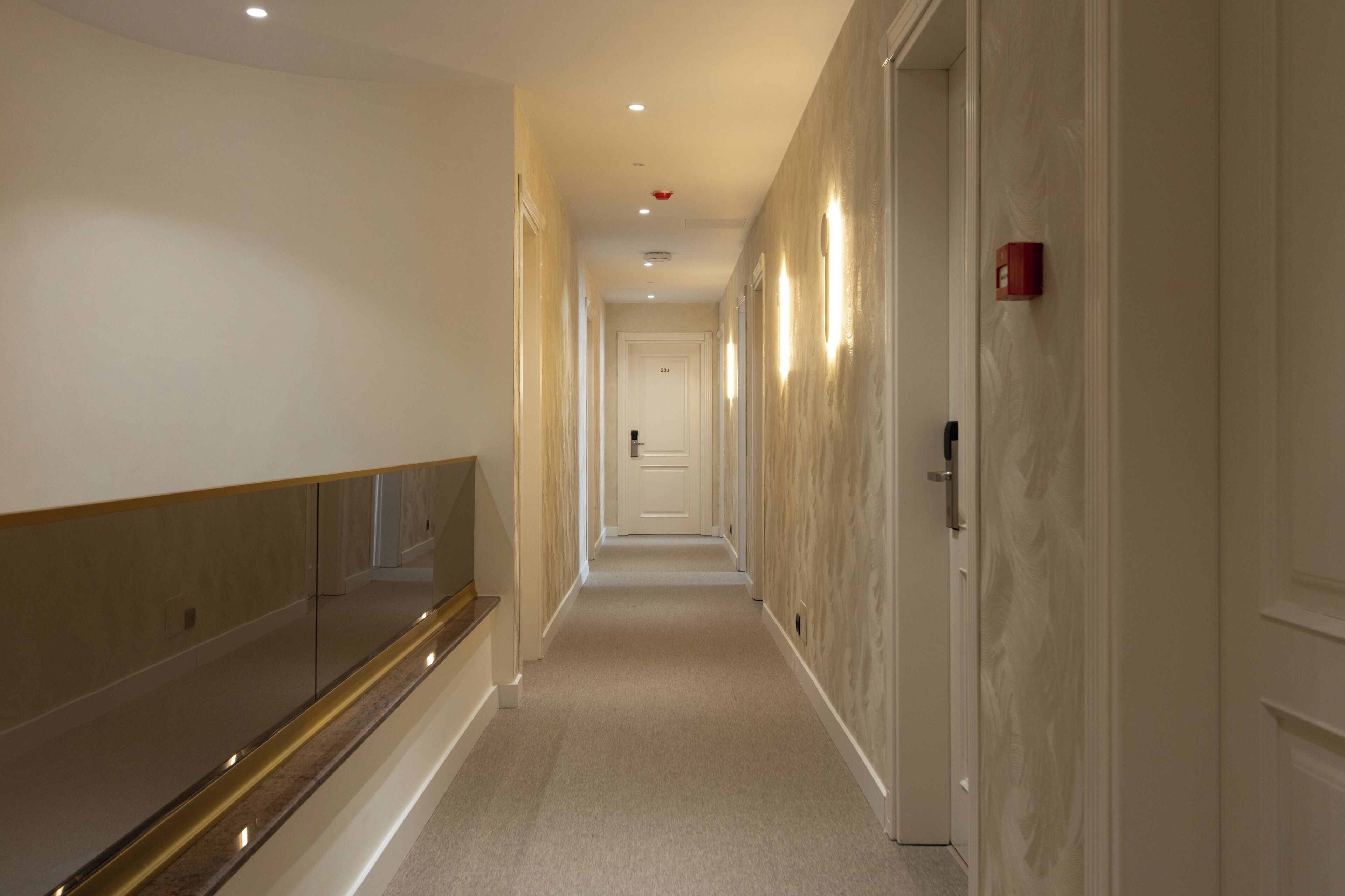 A hotel corridor with a long, narrow layout, a reflective wall, and a door at the end.