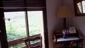 Una habitación con una silla de madera, una mesita con un florero y una ventana con vistas a la vegetación.