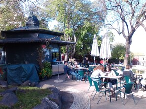 Ein Straßencafé mit Tischen und Stühlen, eine kleine Konstruktion mit Dach und Bäume im Hintergrund.