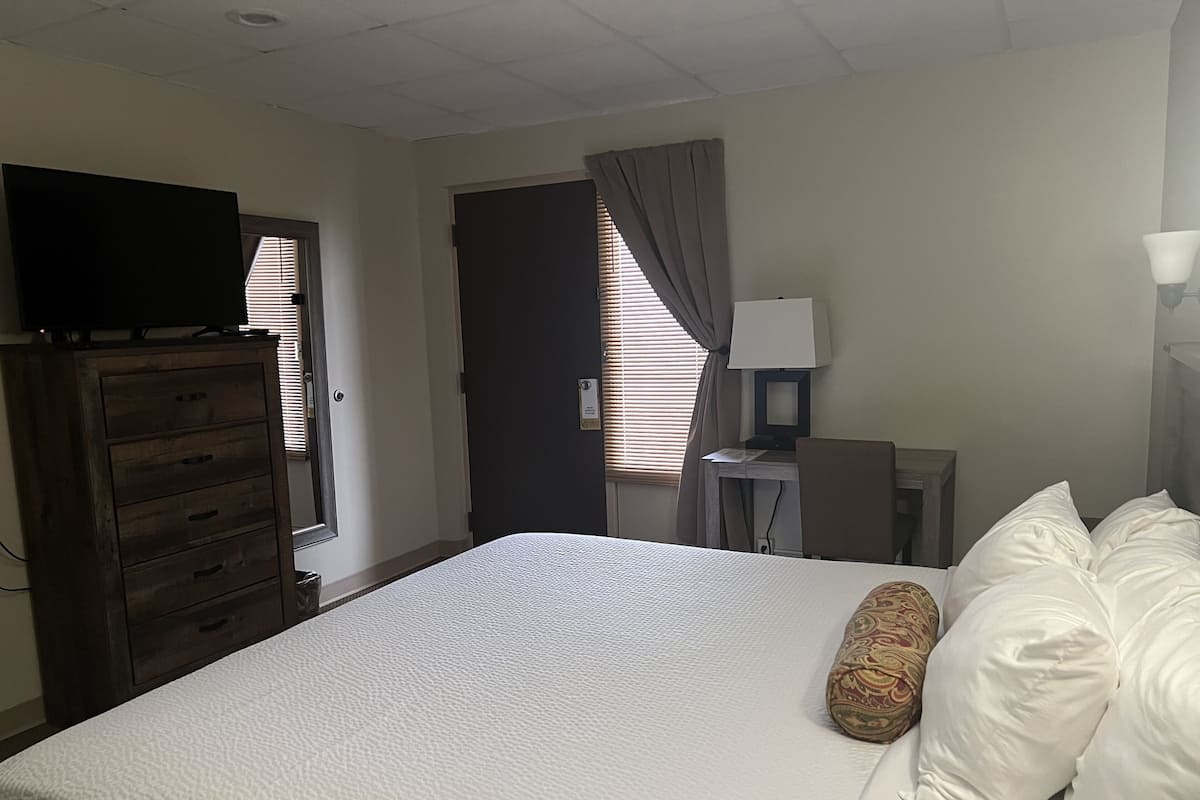 A hotel room with a bed, a desk with a lamp, a television on a dresser, and a window with curtains.