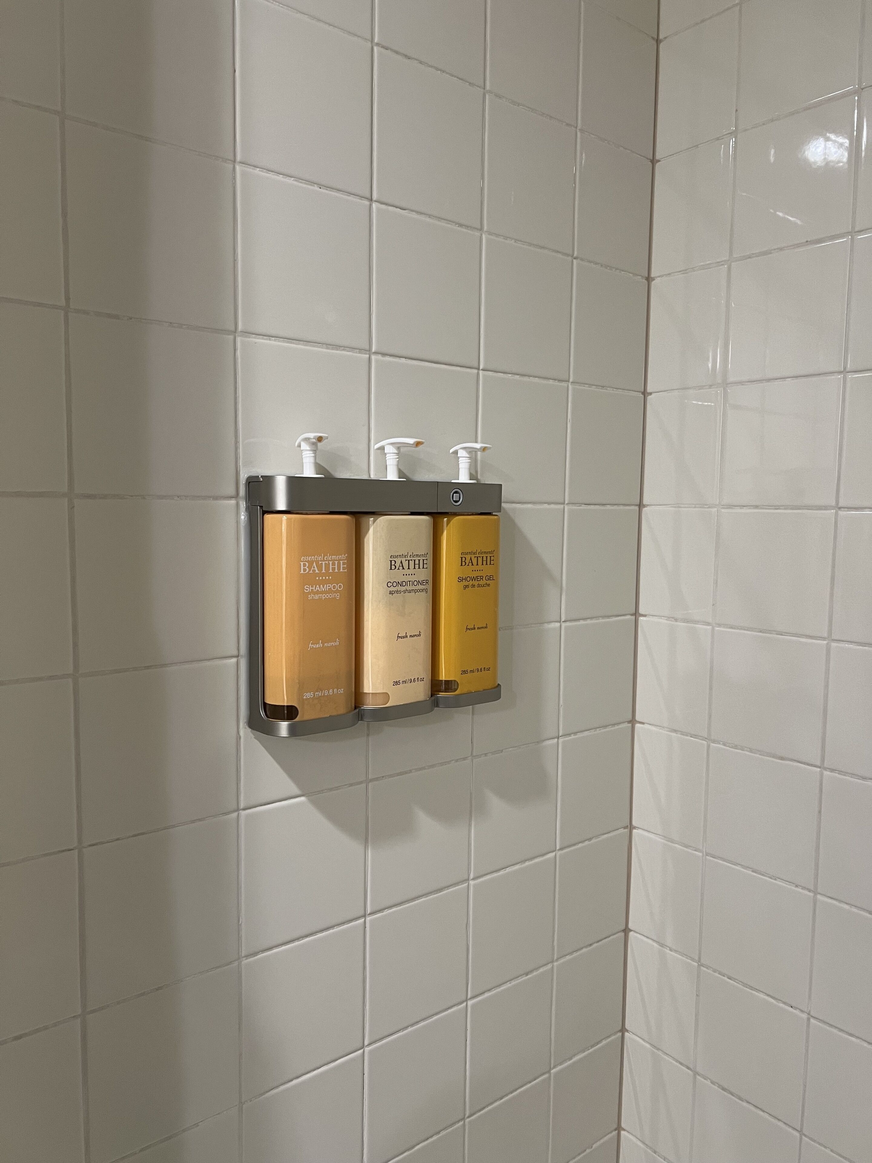 A bathroom with white tiled walls and a wall-mounted dispenser holding three bottles of toiletries.