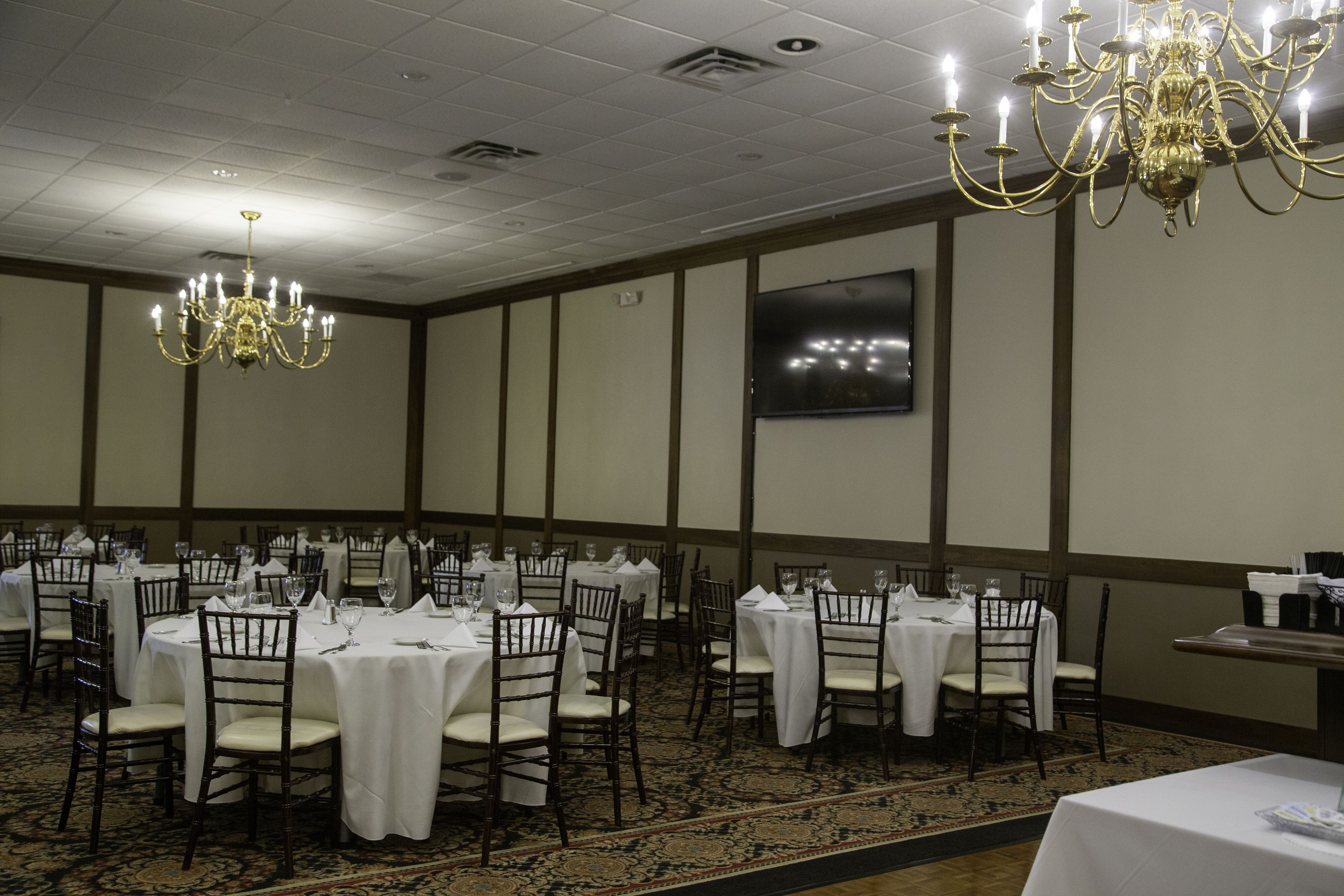 A banquet hall with round tables, chairs, chandeliers, and a large screen on the wall.