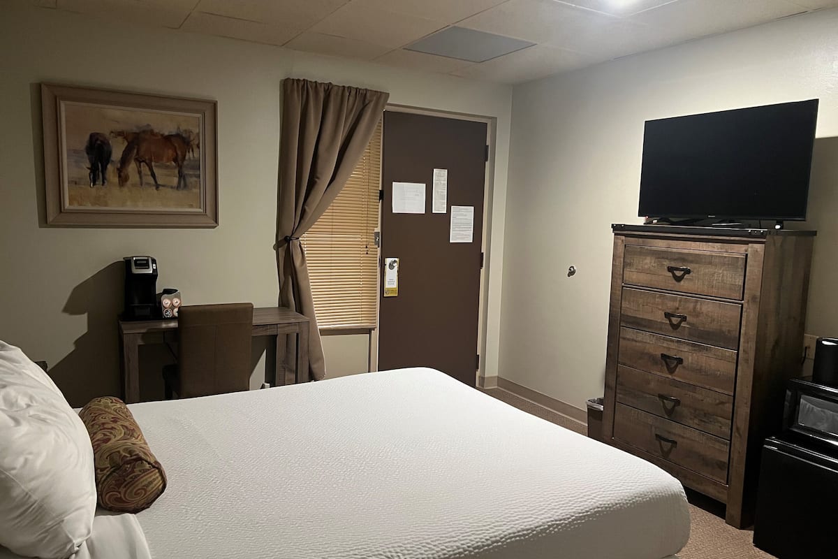 A hotel room with a bed, a desk with a coffee maker, a television on a dresser, and a painting on the wall.