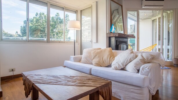 Een moderne woonkamer met een bank, een houten salontafel en grote ramen met uitzicht op bomen.