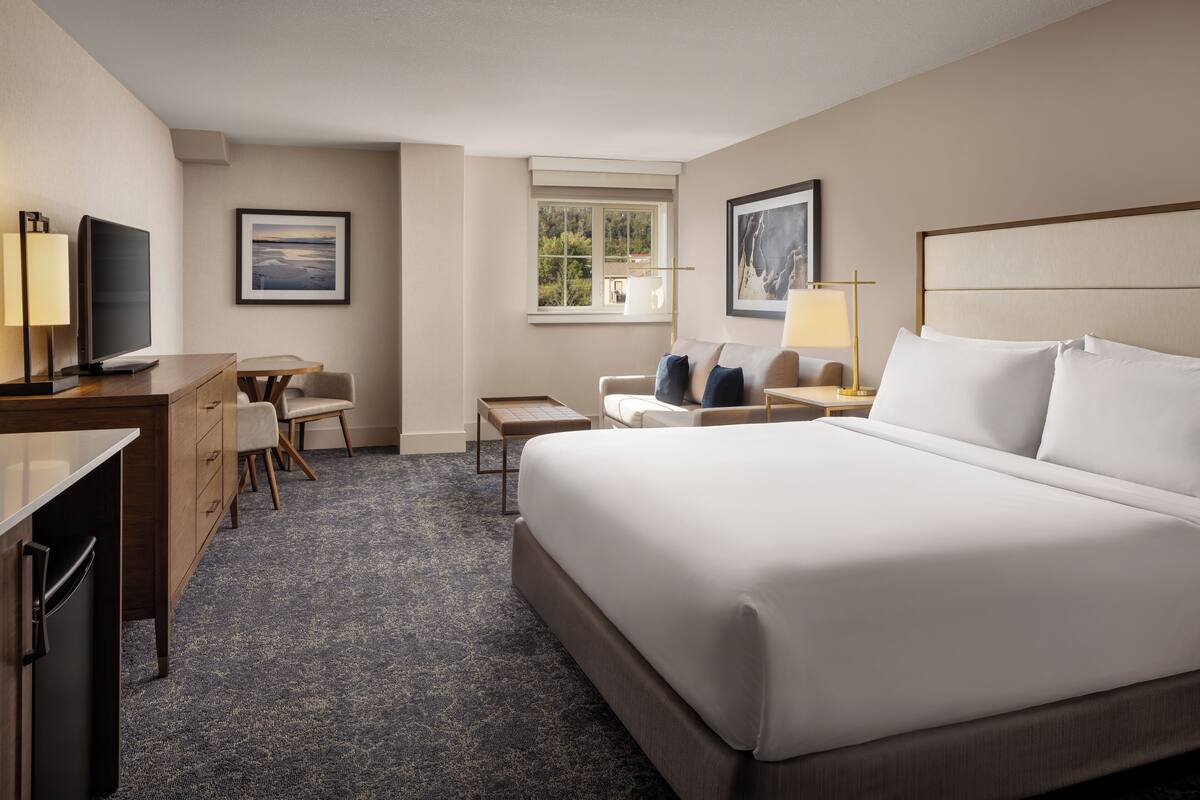A hotel room with a large bed, a desk with a TV, a chair, a small table, and two framed pictures on the walls.
