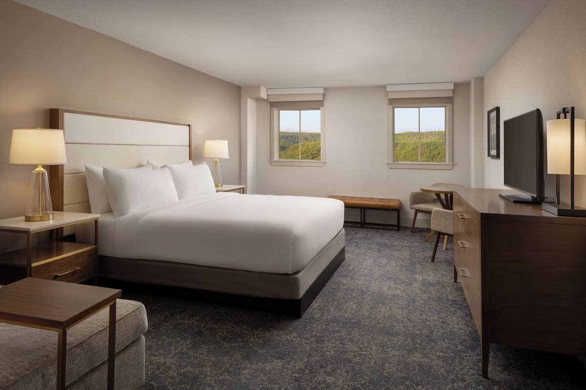 A hotel room with a large bed, bedside tables with lamps, a desk, a chair, and a television.
