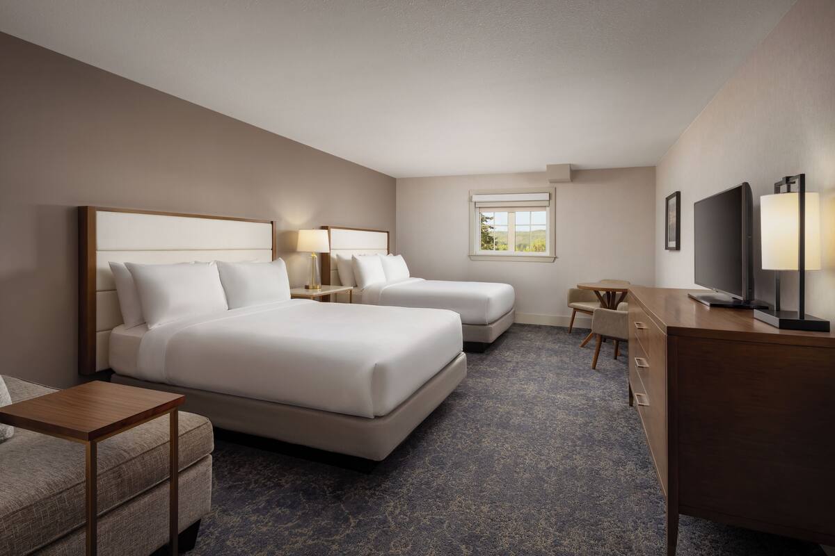 A hotel room with two beds, a desk, a chair, a TV, and a window with a view.