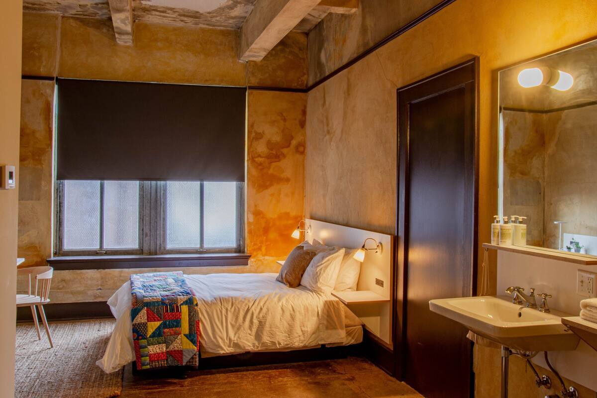 A bedroom with a bed, a bedside table, a window with a view, a bathroom with a sink and mirror.