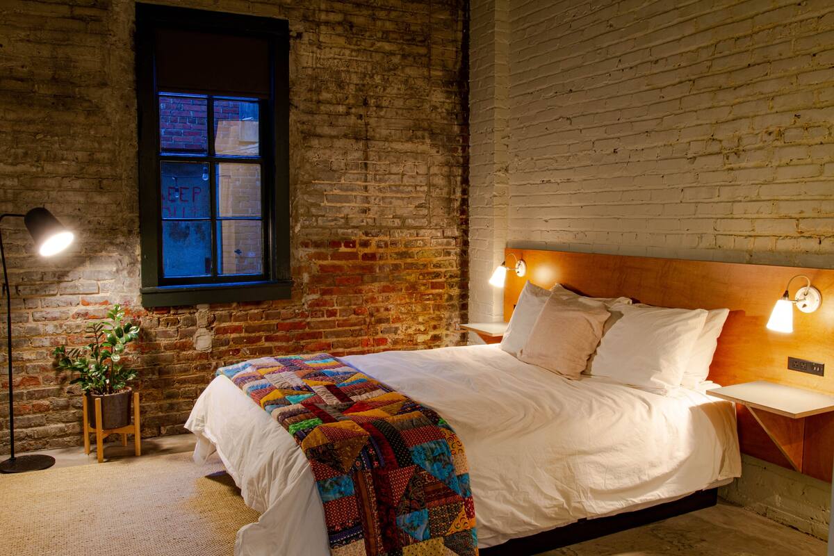 A bedroom with a brick wall, a large window, a bedside table with a lamp, and a bed with a colorful quilt.
