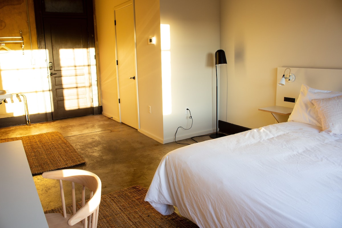 Una habitación de hotel con una cama, un escritorio, una silla, una lámpara y un armario.