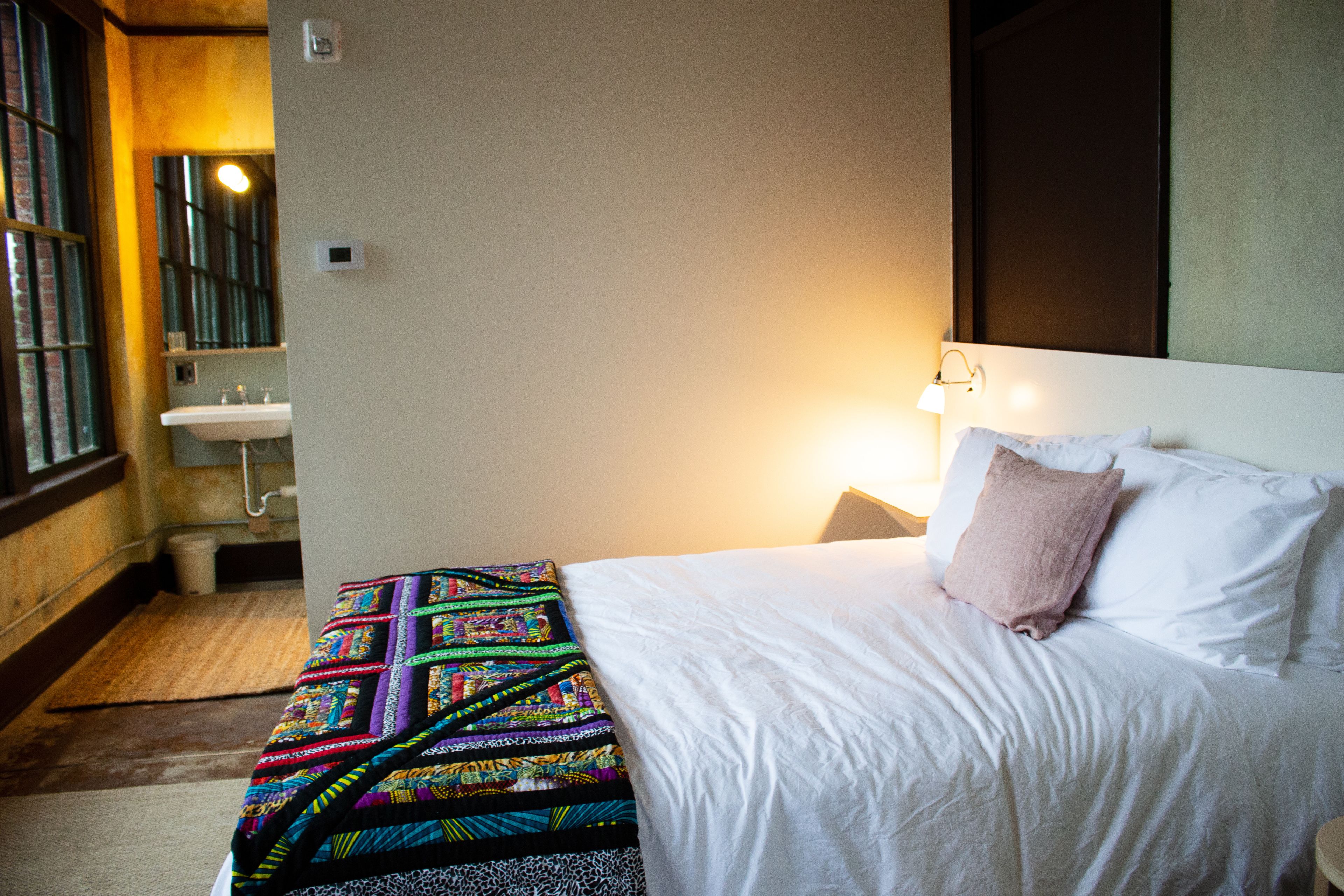 A hotel room with a bed, a bedside table, a nightstand, a window, and a bathroom with a sink and mirror.