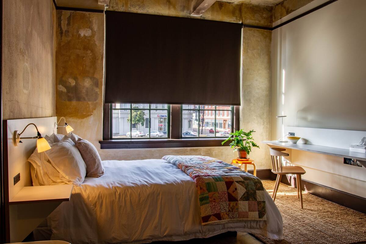 A bedroom with a bed, bedside lamps, a window with a view of the street, a small table with a plant, and a desk with a bowl.