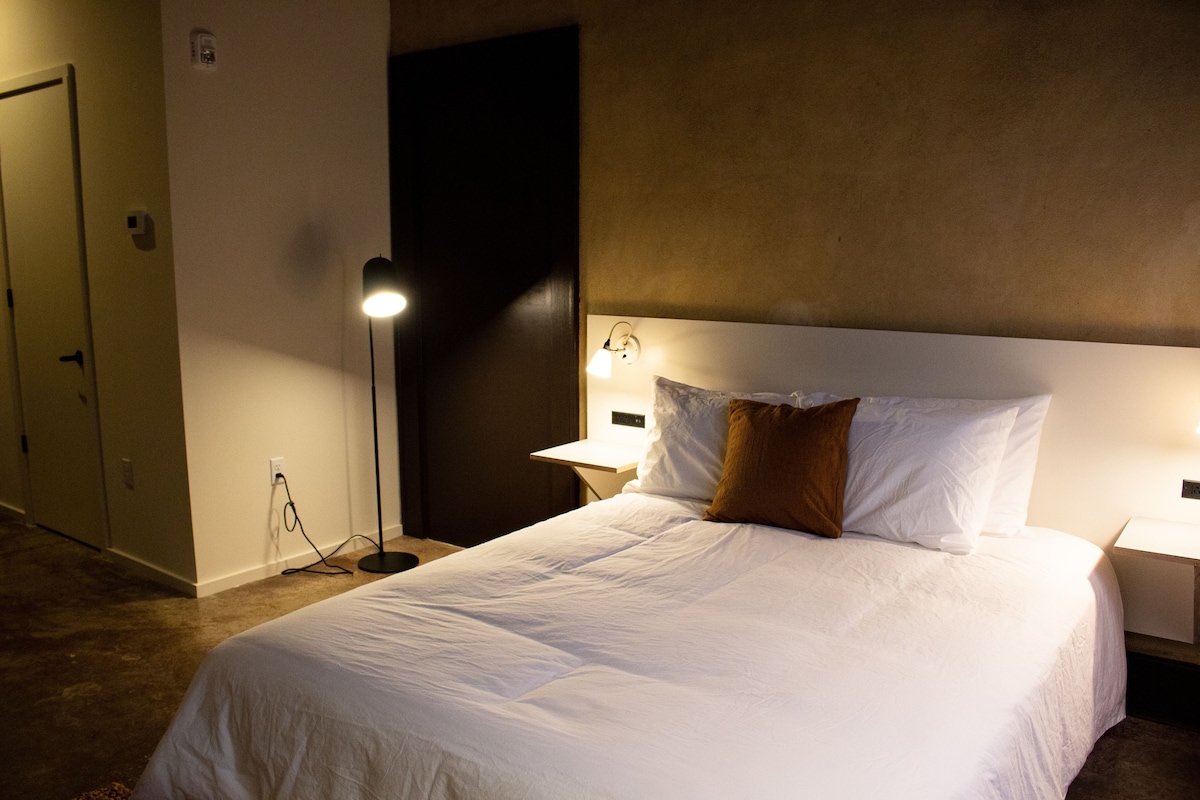 Una habitación de hotel con una cama grande, lámparas de noche, una puerta y una lámpara de pared.